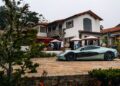 A light green Rimac electric hypercar is parked on a stone driveway in front of a large white house with red roof tiles, surrounded by people and outdoor seating at Monterey Car Week.