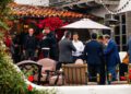 A group of people in formal attire socialize outdoors on a patio with string lights, flowers, and patio furniture during Monterey Car Week, possibly discussing Rimac electric hypercars.