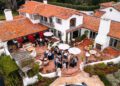 A group of people gathers on a terracotta-tiled patio outside a large white house with a red tile roof; a silver Rimac electric hypercar is parked in the driveway, likely part of the excitement around Monterey Car Week.