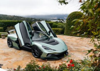 A light green Rimac electric hypercar with its butterfly doors open is parked on a stone driveway surrounded by greenery and flowers, reminiscent of an exclusive Monterey Car Week showcase.