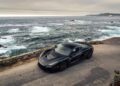 A Rimac electric hypercar is parked on a coastal road beside a stone wall, overlooking the ocean with waves and a rocky shoreline under a cloudy sky—perfect scenery for Monterey Car Week.