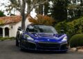 A blue Rimac sports car is parked on a driveway next to a house with a red-tiled roof, surrounded by trees and bushes—an electrifying sight reminiscent of Monterey Car Week.