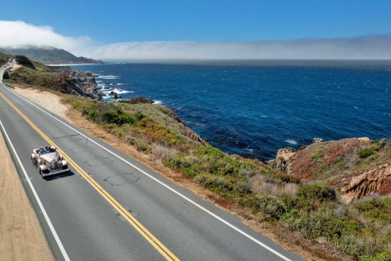 A convertible car cruises the coastal highway under a clear blue sky during Monterey Car Week, cliffs and ocean on one side, hills on the other—capturing the excitement of this famed automotive event.