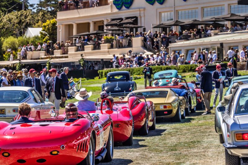 Vintage sports cars are lined up on a lawn at an outdoor car show during Monterey Car Week, with crowds of people and a large building in the background.