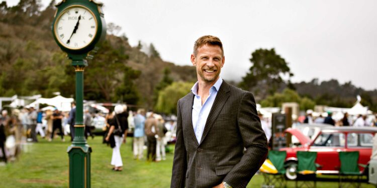 A man in a blazer stands smiling near a green outdoor clock at Rolex Monterey Car Week, with people, cars, and tents in the background on a grassy field.