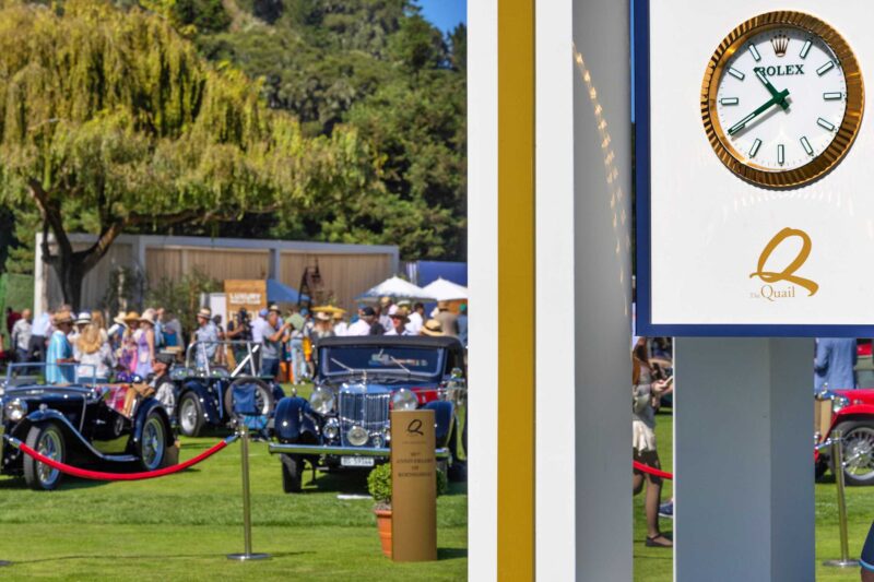 Outdoor classic car show during Monterey Car Week, featuring vintage cars on display, people gathered in the background, and a large Rolex clock in the foreground showing the time.