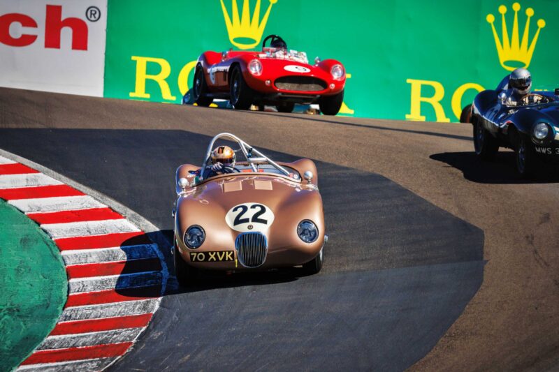 Three vintage race cars speed around a curved section of the track at Monterey Car Week, with car number 22 leading the pack in this thrilling showcase of classic racing.