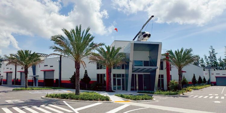 Modern commercial building with palm trees, a glass entrance, and a helicopter mounted on the roof, featuring state-of-the-art storage under a partly cloudy sky.