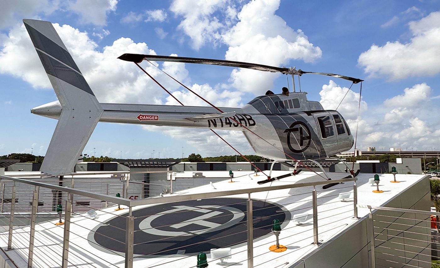 A silver helicopter with the registration N7149B is displayed on a rooftop helipad at The Hangar, anchored with red straps under a partly cloudy sky.