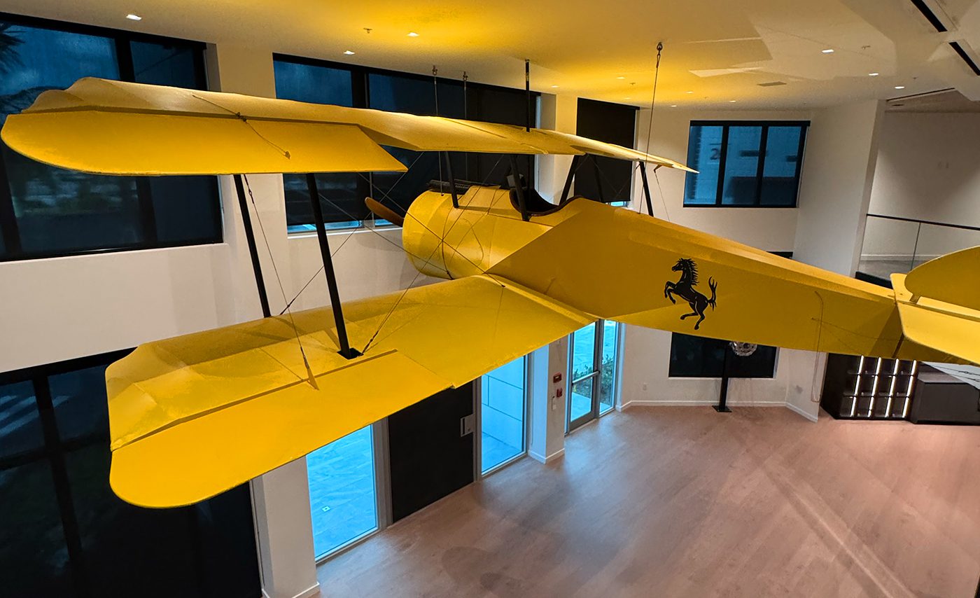 A large yellow biplane model with a black prancing horse logo is suspended from the ceiling in The Hangar, a modern, open indoor space with wood flooring, large windows, and state-of-the-art storage.