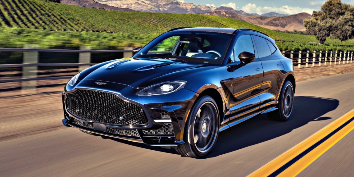 A black 2026 Aston Martin DBX S First Drive: The Best Super SUV Gets Even Better cruises on a paved road past a vineyard, framed by mountains and blue sky.
