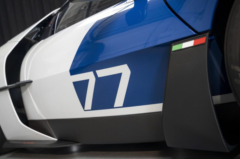 Close-up of a blue and white race car with the number 77 on the side and a small Italian flag decal above a carbon fiber side panel.