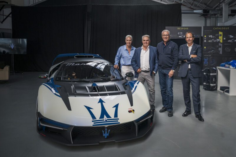 Four men stand next to a white and blue Maserati sports car inside an indoor showroom with automotive displays in the background.