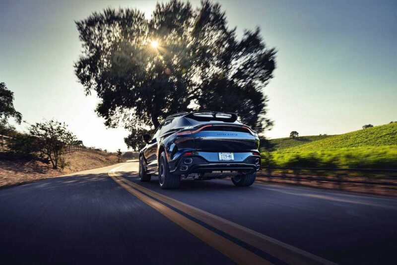 The 2026 Aston Martin DBX S First Drive: The Best Super SUV Gets Even Better as it cruises along a country road bordered by trees and grassy fields, sunlight streaming through the branches ahead.