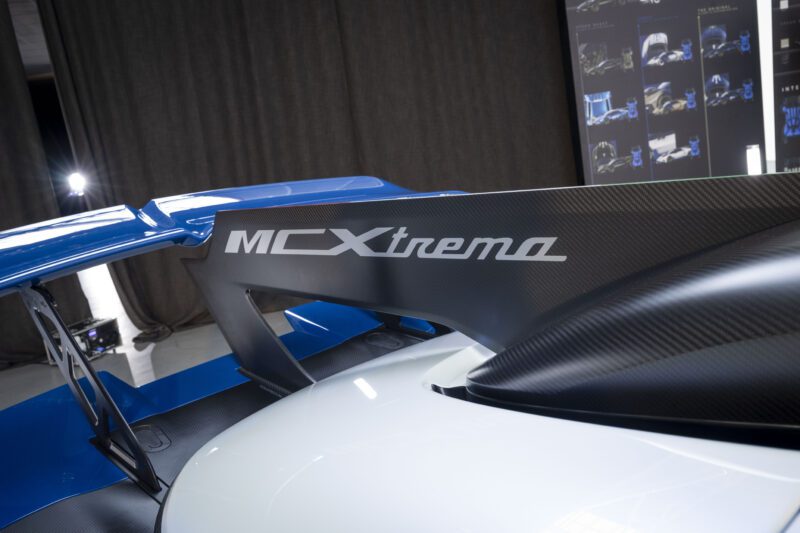 Close-up of a car’s rear with a large carbon fiber wing labeled “MCXtrema” and a blue spoiler; design sketches are visible in the blurred background.