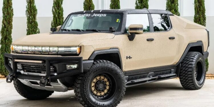 A tan custom Hummer EV pickup truck with off-road tires, a unique creation among custom cars for sale, is parked on concrete in front of tall, trimmed evergreen trees near a garage.