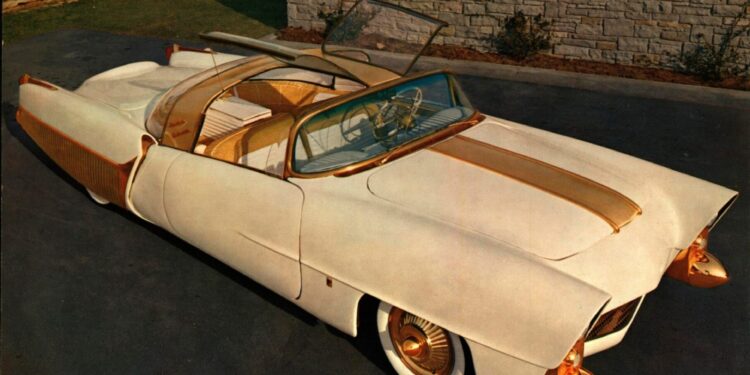 The Golden Sahara II, a vintage white and gold convertible with a transparent roof, is parked on a driveway near a stone wall.