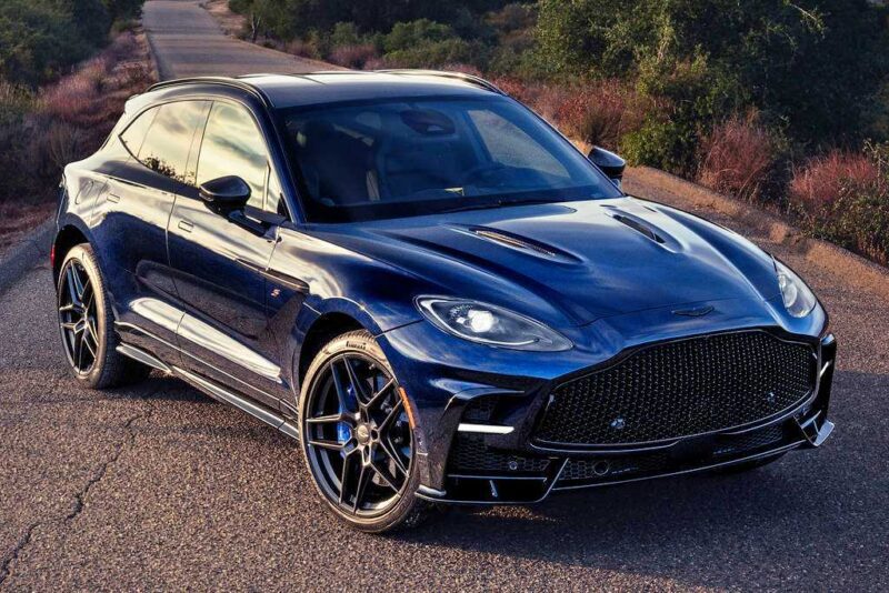 A dark blue 2026 Aston Martin DBX S First Drive: The Best Super SUV Gets Even Better, parked on a rural road surrounded by dry grass and trees, photographed in daylight.