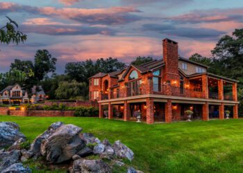 A large two-story brick house with a wraparound patio and outdoor lighting sits on a green lawn at sunset, offering the charm of a mountain-view country estate, with trees and another house in the background.