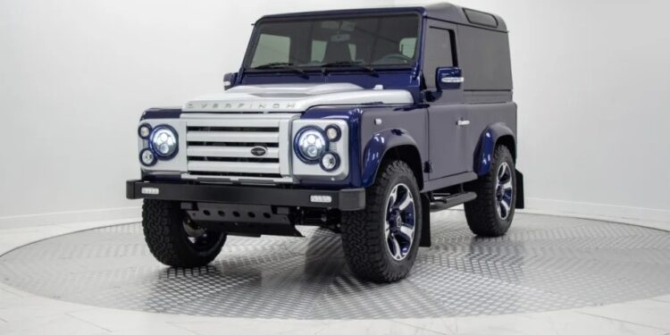 A blue and silver Land Rover Defender, a unique creation with customized features, is parked in a white, modern indoor showroom on a circular display platform.