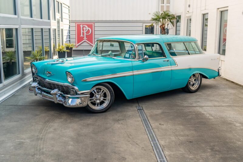 A turquoise and white 1956 Chevrolet Bel Air Nomad station wagon, a standout among classic muscle cars, is parked in a courtyard with glass walls and potted plants.