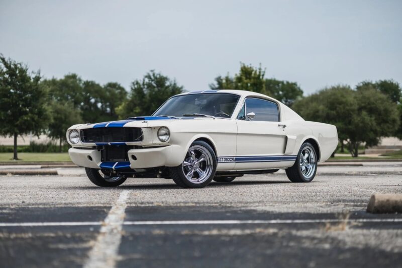 A classic white Ford Mustang fastback with blue racing stripes, a true gem among Classic Muscle Cars, is parked in an empty parking lot with trees in the background.