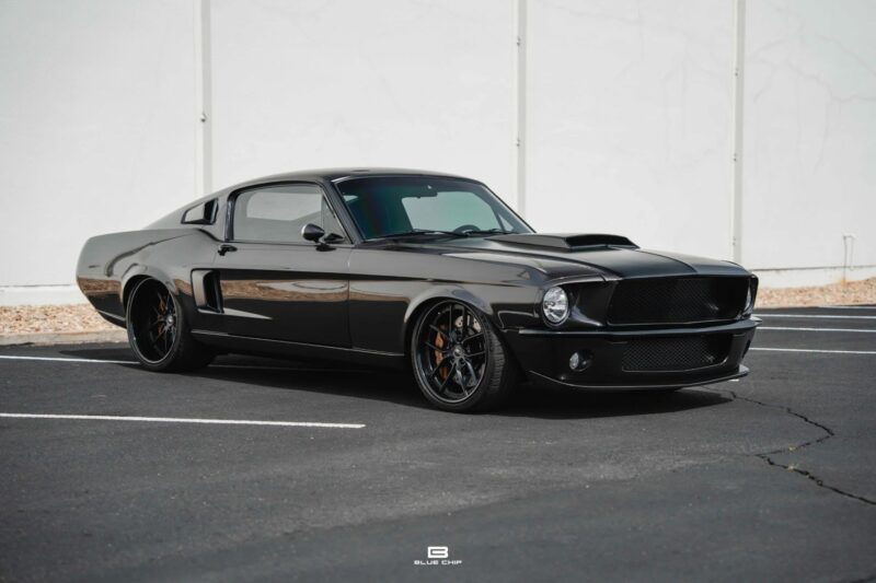 A black classic muscle car, a true icon among Classic Muscle Cars, is parked in an empty lot with a white wall in the background.