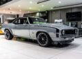 A silver 1969 Chevrolet Camaro SS coupe, a true icon among Muscle Cars, is displayed indoors with black racing stripes, large black wheels, and a polished showroom environment.