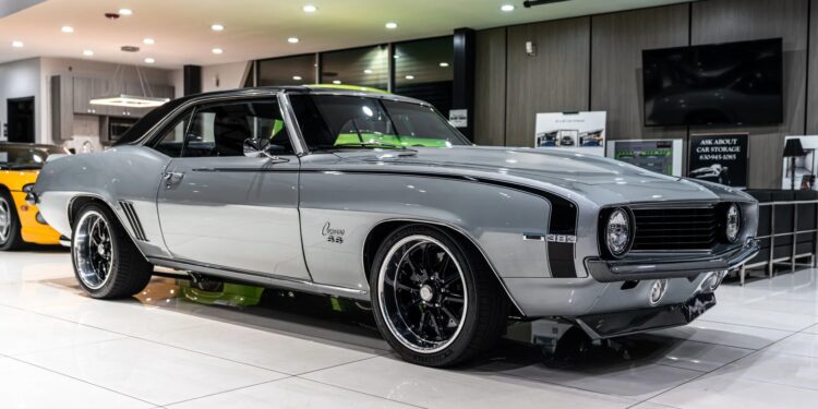 A silver 1969 Chevrolet Camaro, a standout among classic muscle cars, with black rims is displayed indoors under bright lights on a glossy white floor.