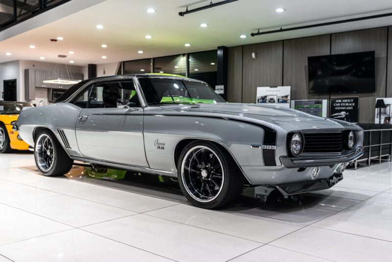 A silver 1969 Chevrolet Camaro, a standout among classic muscle cars, with black rims is displayed indoors under bright lights on a glossy white floor.