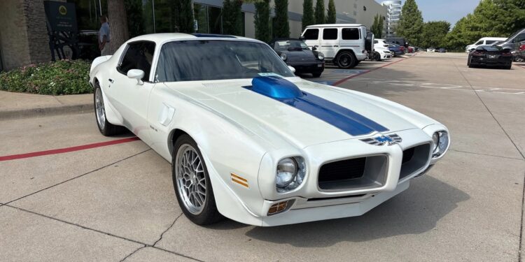 A white classic muscle car with a blue stripe and hood scoop is parked in a lot near a modern building and other vehicles—a standout among muscle cars and perfect for anyone searching classic cars for sale.