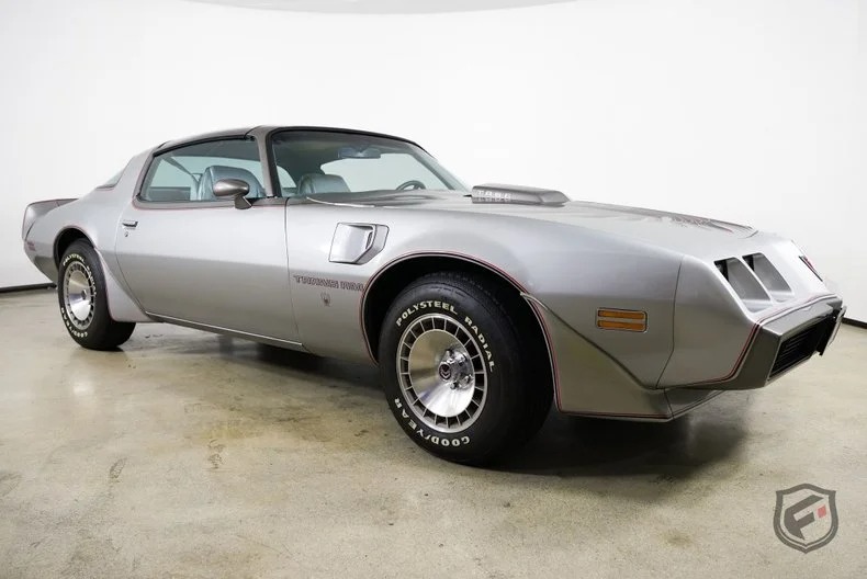 A silver classic Pontiac Trans Am coupe, a standout among classic muscle cars, is parked indoors on a polished concrete floor, viewed from the front driver's side.