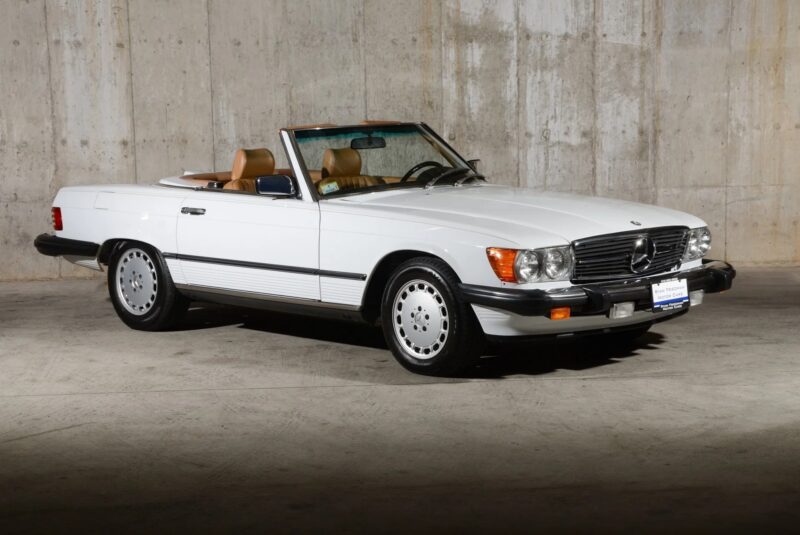 This luxury car, a white Mercedes-Benz convertible with tan interior, is parked indoors against a concrete wall and angled slightly to the left.
