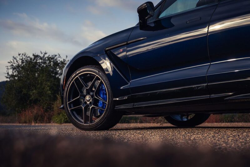Close-up of the front side of a dark-colored 2026 Aston Martin DBX S First Drive: The Best Super SUV Gets Even Better, parked on a road, revealing the wheel, brake caliper, and part of the hood under natural light.