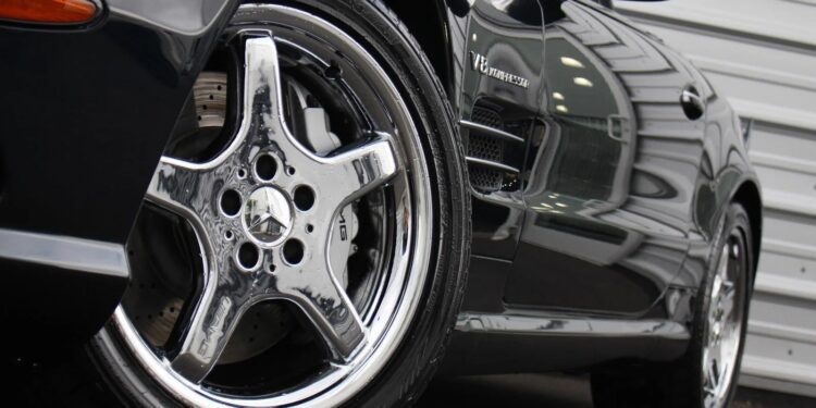 Close-up view of a luxury Mercedes-Benz sports car, focusing on a shiny chrome wheel and part of the front fender, parked near a metal-clad building.