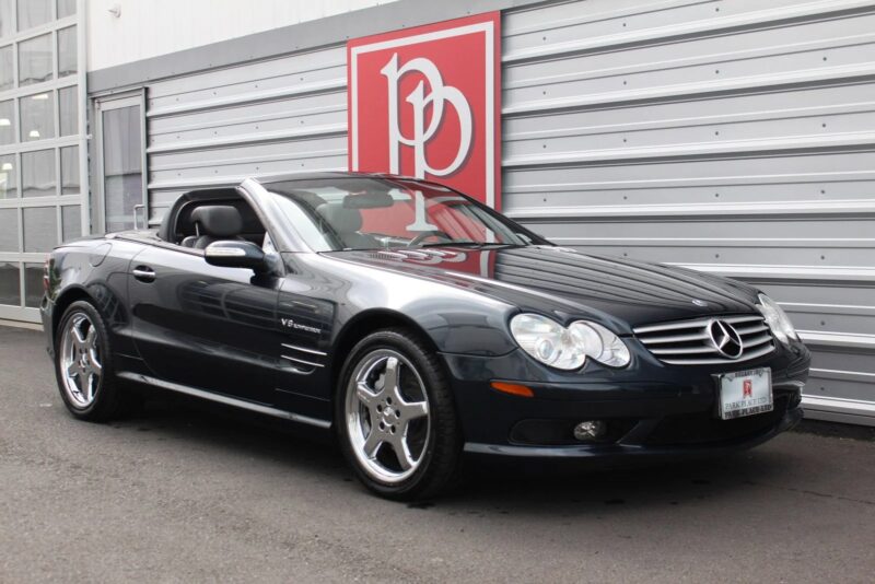 A luxury car, this black Mercedes-Benz SL55 AMG convertible is parked in front of a building with a large red and white sign, ready for sale.