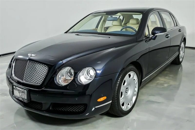A dark blue Bentley sedan, a standout among luxury sedans for sale, is parked in a showroom with its cream-colored interior visible through the windows.
