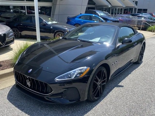 A black Maserati GranTurismo convertible, a true luxury car, is parked on a paved lot near a car dealership, with other vehicles visible in the background.