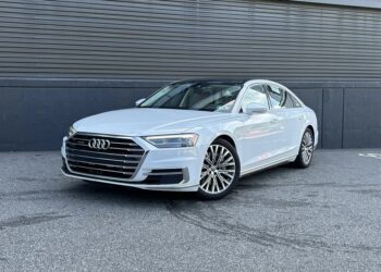 A white Audi sedan, a standout among affordable luxury sedans, is parked on a paved surface in front of a dark, ribbed metal wall.