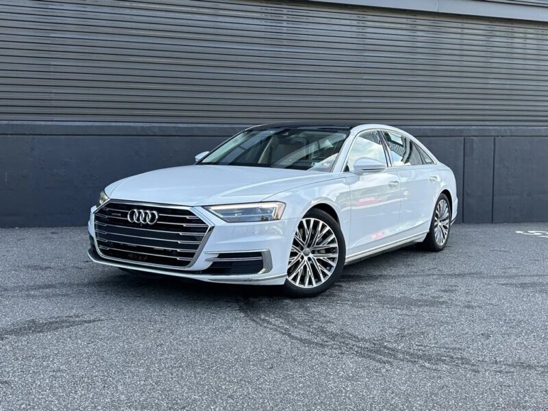 A white Audi sedan, a standout among affordable luxury sedans, is parked on a paved surface in front of a dark, ribbed metal wall.