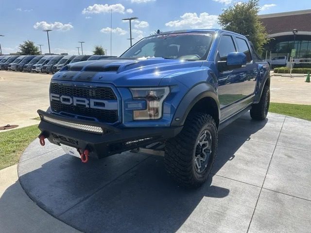 A blue Ford F-150 Shelby pickup with off-road tires and a custom front bumper is parked on a concrete platform outdoors under a sunny sky.