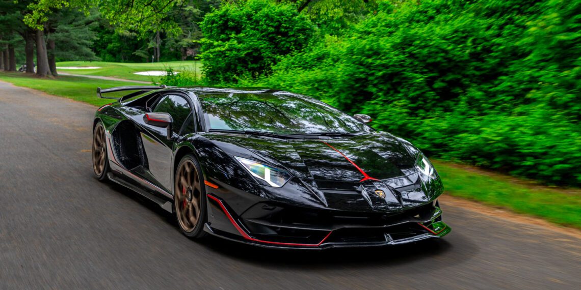 A black Lamborghini sports car with red accents drives on a tree-lined road, surrounded by green foliage and grass.