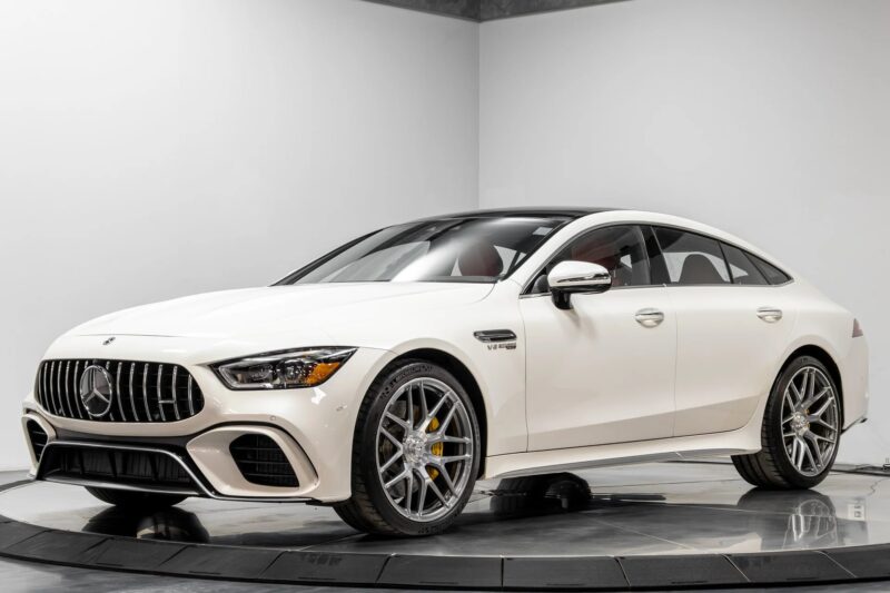 A white Mercedes-Benz AMG GT four-door luxury car is displayed in a showroom, viewed from the front left side.