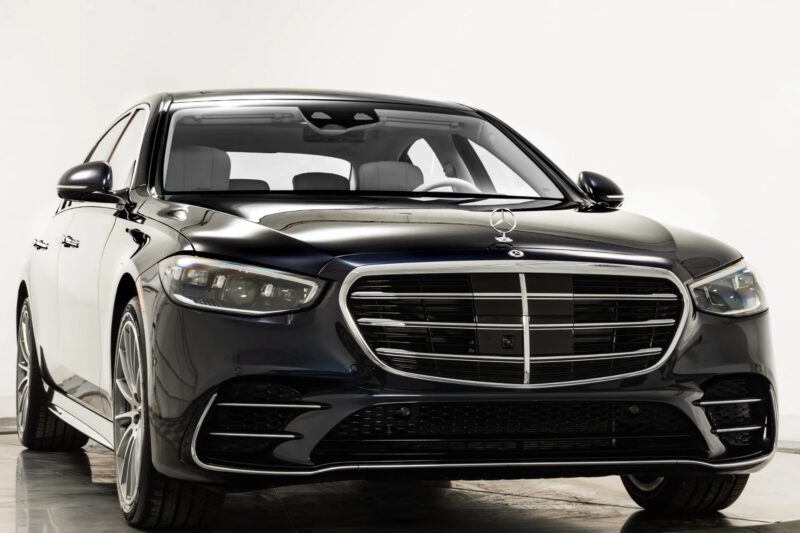 A black Mercedes-Benz sedan, one of the top affordable luxury sedans, is parked indoors on a reflective surface, shown from a front angle against a plain white background.