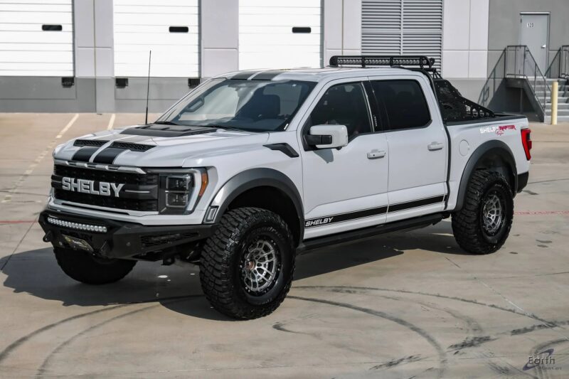 A white Ford F-150 Shelby, one of the best Ford trucks, with black stripes, raised suspension, off-road tires, and aftermarket accessories is parked on concrete near industrial buildings.