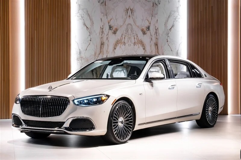 A white Mercedes-Benz luxury sedan with chrome accents and intricate alloy wheels is displayed indoors under bright lights, positioned in front of a marble and wood backdrop.