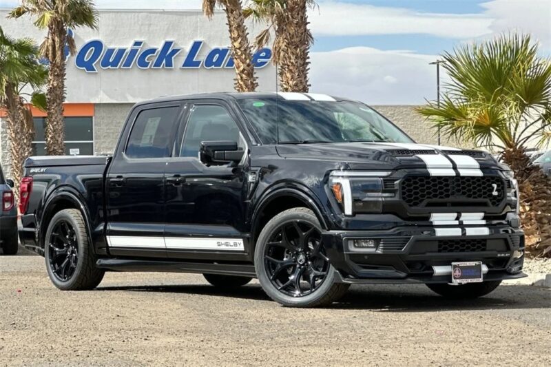 A black Ford F-150 Shelby pickup truck with white racing stripes is parked outdoors near palm trees and a Quick Lane service center. Discover the power of Shelby Trucks—your next Ford F-150 for sale could be here.