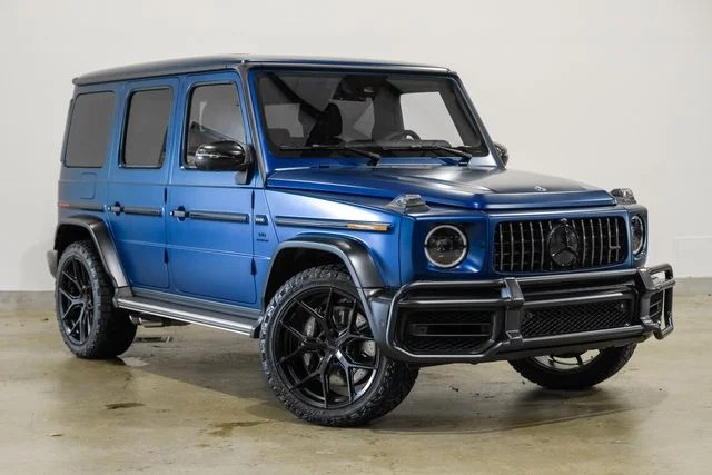 A blue Mercedes-Benz G-Class luxury car with black rims is parked indoors on a concrete floor against a plain white background.