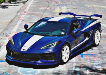 A blue Yenko C8 Corvette sports car with white racing stripes and a large rear spoiler is parked on a graffiti-covered road.