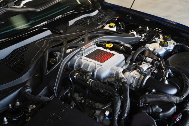 Vista de cerca del compartimiento del motor de un automóvil con supercargador visible, mangueras y componentes mecánicos de First Drive: el Ford Mustang GTD de seis cifras presume de su máximo músculo pony.
